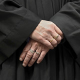 Zu sehen sind zwei Hände einer männlichen Person, vor sich verschränkt. Die Person trägt einen Talar.